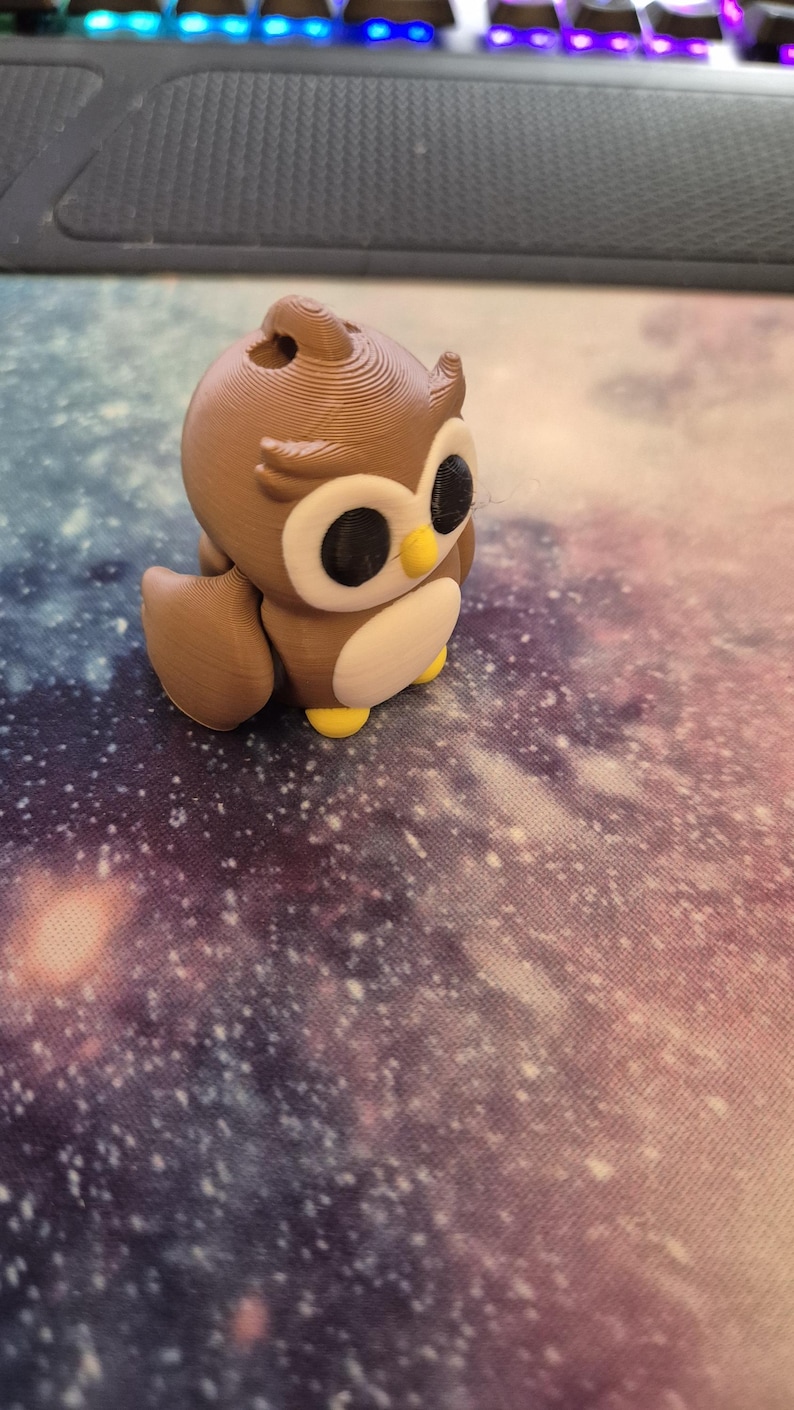May include: A brown and white owl figurine with large black eyes. The owl is sitting on a purple and white galaxy-themed surface.