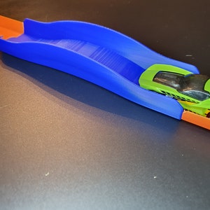 Op de afbeelding: Een blauw plastic baanstuk met een groene speelgoedauto die eroverheen rijdt. Het baanstuk is gebogen en heeft een lichte helling.