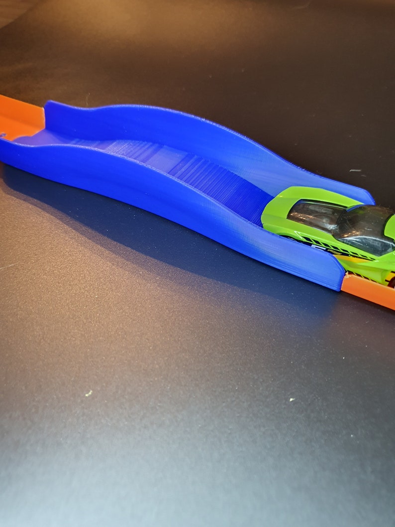 Op de afbeelding: Een blauw plastic baanstuk met een groene speelgoedauto die eroverheen rijdt. Het baanstuk is gebogen en heeft een lichte helling.