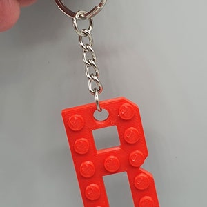 Peut inclure: Un porte-cl&eacute;s en plastique rouge en forme de lettre R, con&ccedil;u pour ressembler &agrave; une brique Lego. Le porte-cl&eacute;s est attach&eacute; &agrave; un anneau et une cha&icirc;ne en m&eacute;tal argent&eacute;.