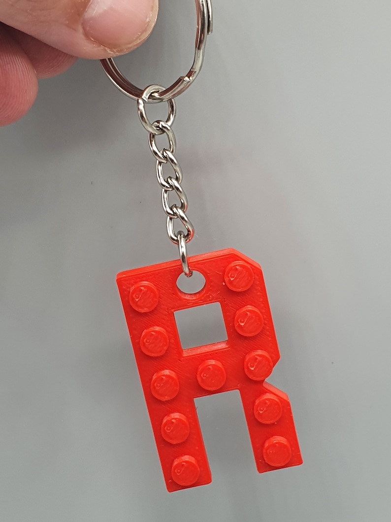 Peut inclure: Un porte-cl&eacute;s en plastique rouge en forme de lettre R, con&ccedil;u pour ressembler &agrave; une brique Lego. Le porte-cl&eacute;s est attach&eacute; &agrave; un anneau et une cha&icirc;ne en m&eacute;tal argent&eacute;.