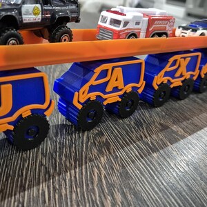 May include: A toy truck carrying a line of smaller toy trucks, each with a letter of the alphabet on the side. The trucks are blue and orange and are arranged in alphabetical order, starting with J and ending with N.