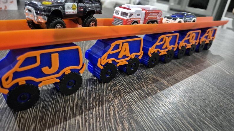 May include: A toy truck carrying a line of smaller toy trucks, each with a letter of the alphabet on the side. The trucks are blue and orange and are arranged in alphabetical order, starting with J and ending with N.