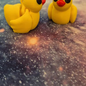May include: Two yellow rubber duck figurines with black eyes and red beaks. The ducks are sitting on a galaxy-patterned surface.