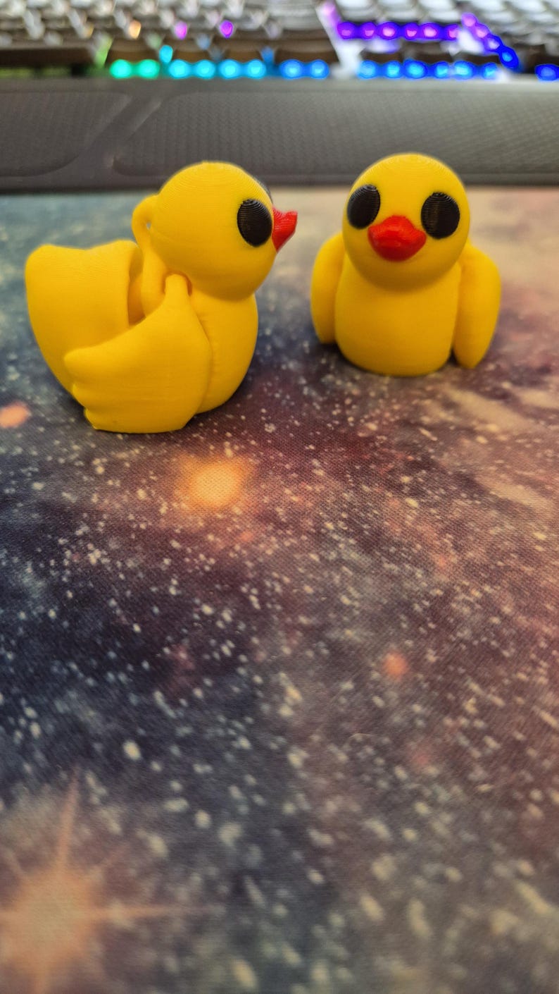 May include: Two yellow rubber duck figurines with black eyes and red beaks. The ducks are sitting on a galaxy-patterned surface.
