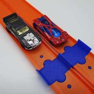 K&ouml;nnte beinhalten: Zwei Spielzeugautos fahren auf einer orangefarbenen Rennstrecke mit einer blauen Plastikrampe. Das Auto links ist schwarz mit einem silbernen Motor und das Auto rechts ist rot mit einem Spinnennetz-Design.