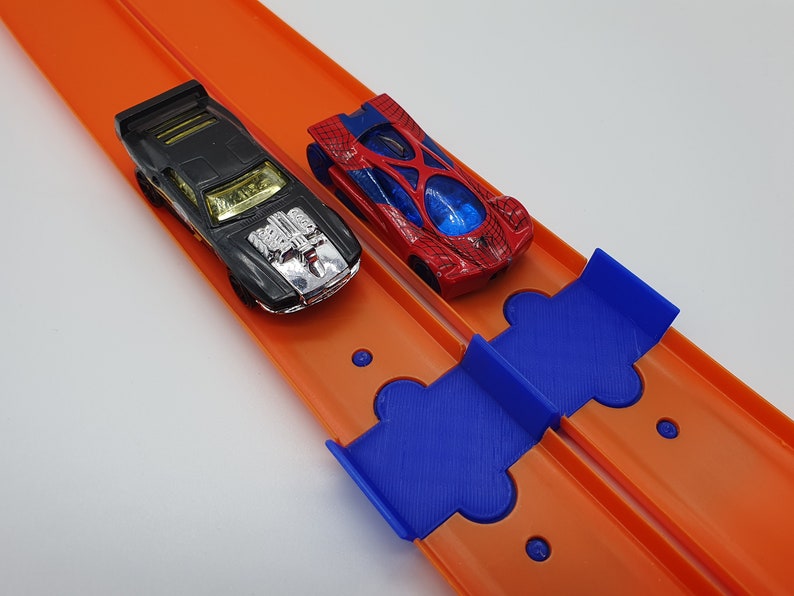 K&ouml;nnte beinhalten: Zwei Spielzeugautos fahren auf einer orangefarbenen Rennstrecke mit einer blauen Plastikrampe. Das Auto links ist schwarz mit einem silbernen Motor und das Auto rechts ist rot mit einem Spinnennetz-Design.
