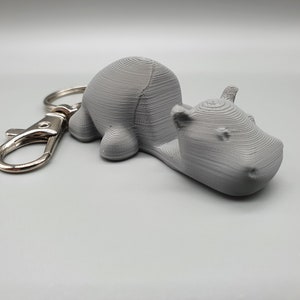 May include: A grey 3D printed hippopotamus keychain with a silver metal key ring.