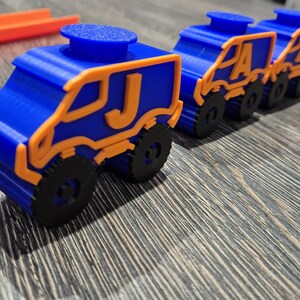 May include: A line of blue toy trucks with orange accents and black wheels. Each truck has a letter on the side, starting with the letter J.