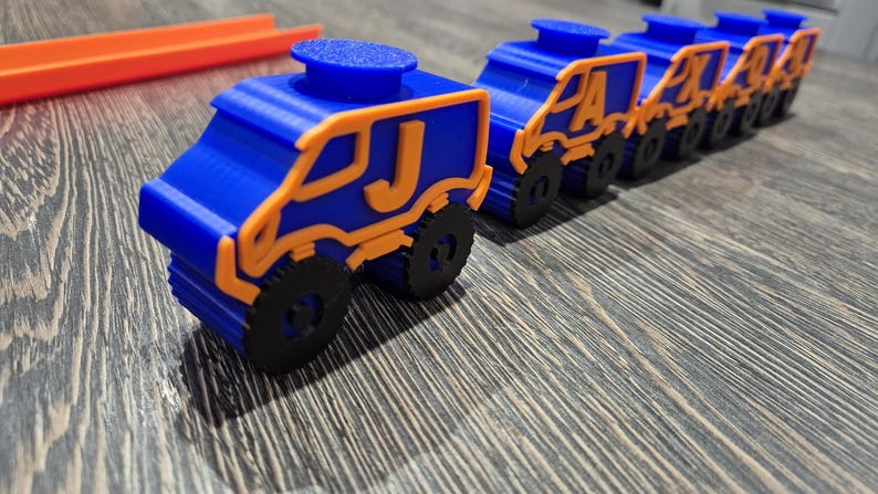 May include: A line of blue toy trucks with orange accents and black wheels. Each truck has a letter on the side, starting with the letter J.