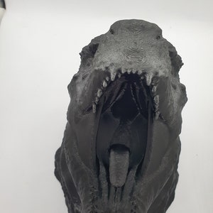 May include: A black 3D printed dinosaur head with an open mouth and a visible tongue. The dinosaur head is facing the camera.