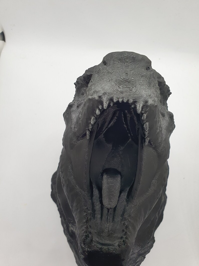 May include: A black 3D printed dinosaur head with an open mouth and a visible tongue. The dinosaur head is facing the camera.