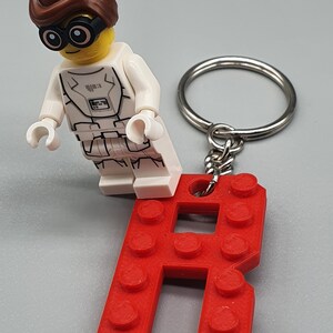 Peut inclure: Un porte-cl&eacute;s en forme de lettre R en Lego rouge avec une figurine Lego blanche portant une combinaison spatiale blanche et des cheveux bruns.