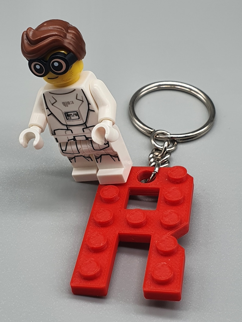 Peut inclure: Un porte-cl&eacute;s en forme de lettre R en Lego rouge avec une figurine Lego blanche portant une combinaison spatiale blanche et des cheveux bruns.