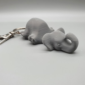 May include: A gray 3D printed elephant keychain with a smaller elephant attached to the larger one. The keychain has a silver metal ring.