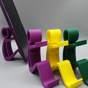 May include: Four phone stands in purple, yellow, and green. The stands are shaped like stick figures sitting with their legs bent. The stands are designed to hold a phone upright.