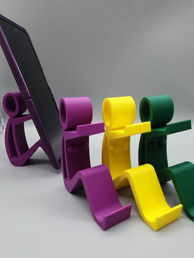 May include: Four phone stands in purple, yellow, and green. The stands are shaped like stick figures sitting with their legs bent. The stands are designed to hold a phone upright.