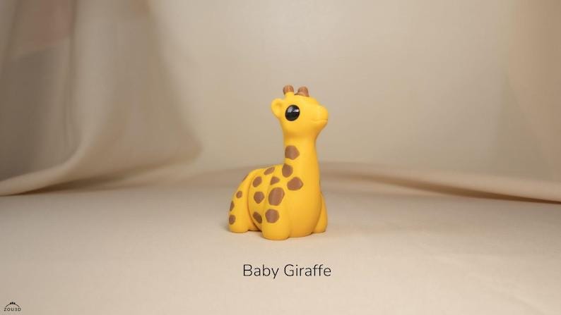 May include: A yellow baby giraffe figurine with brown spots. The giraffe is sitting down with its head up, and has large black eyes. The text "Baby Giraffe" is below the figurine.