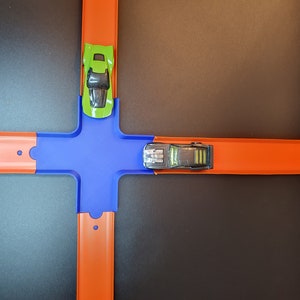 May include: A toy car track set with two toy cars. One car is green and the other is black. The track is orange and blue.