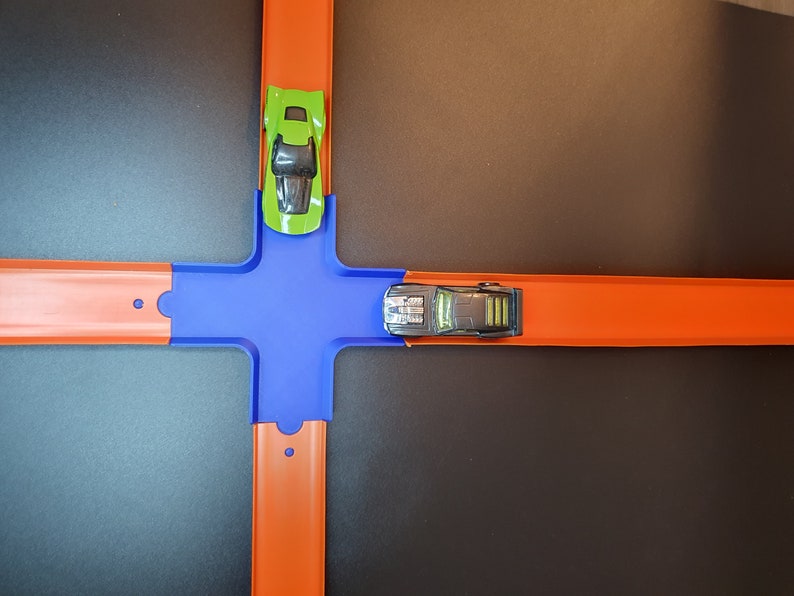 May include: A toy car track set with two toy cars. One car is green and the other is black. The track is orange and blue.