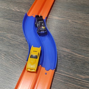 May include: A blue and orange toy race track with two toy cars. One car is yellow and the other is black with a police car design. The cars are on the track and ready to race.