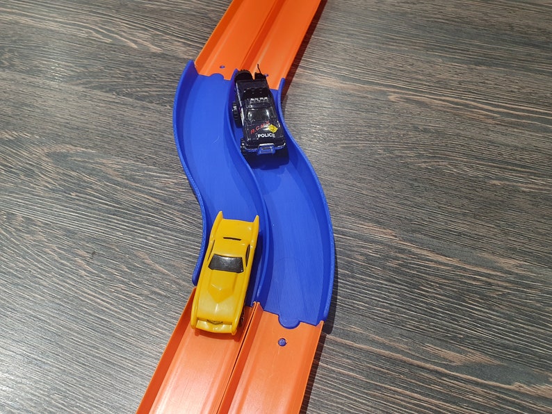 May include: A blue and orange toy race track with two toy cars. One car is yellow and the other is black with a police car design. The cars are on the track and ready to race.