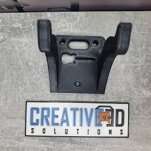 May include: A black 3D printed wall mount for a guitar. The mount has a cutout for the guitar's headstock and a hole for mounting screws. The mount is labeled "CREATIVE 3D SOLUTIONS".