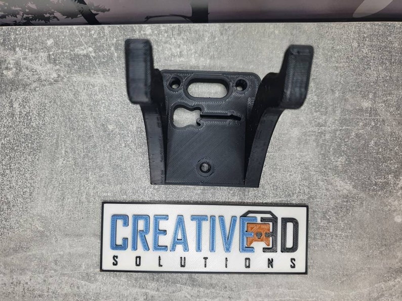 May include: A black 3D printed wall mount for a guitar. The mount has a cutout for the guitar's headstock and a hole for mounting screws. The mount is labeled "CREATIVE 3D SOLUTIONS".