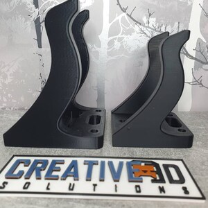 May include: Two black 3D printed wall mounts with a curved design. The mounts are designed to hold a device or object. The mounts are made by Creative 3D Solutions.