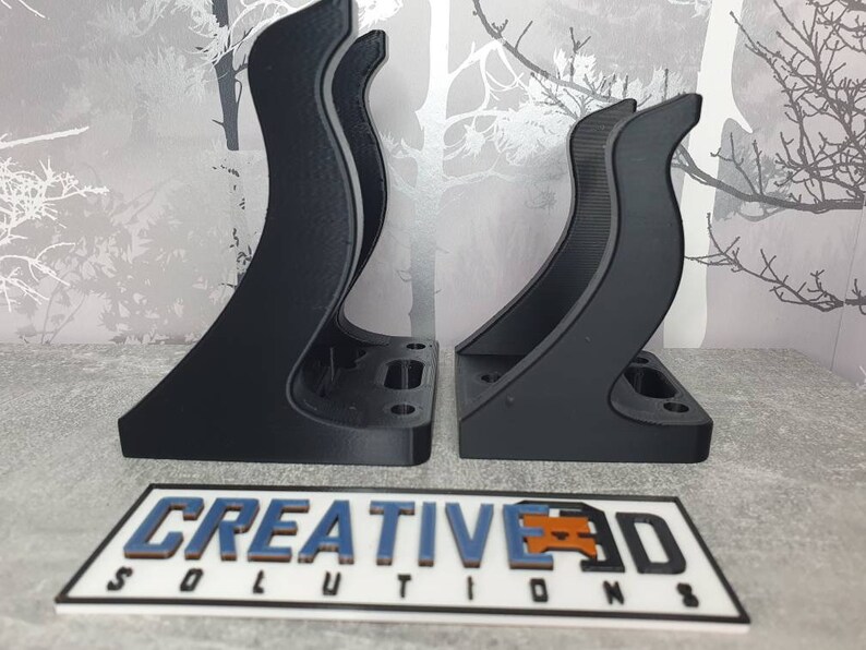 May include: Two black 3D printed wall mounts with a curved design. The mounts are designed to hold a device or object. The mounts are made by Creative 3D Solutions.