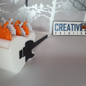 May include: A white 3D printed guitar with a black neck and a row of orange and white pixelated space invaders on top. The guitar is sitting on a gray surface. The text "CREATIVE 3D SOLUTIONS" is visible in the background.