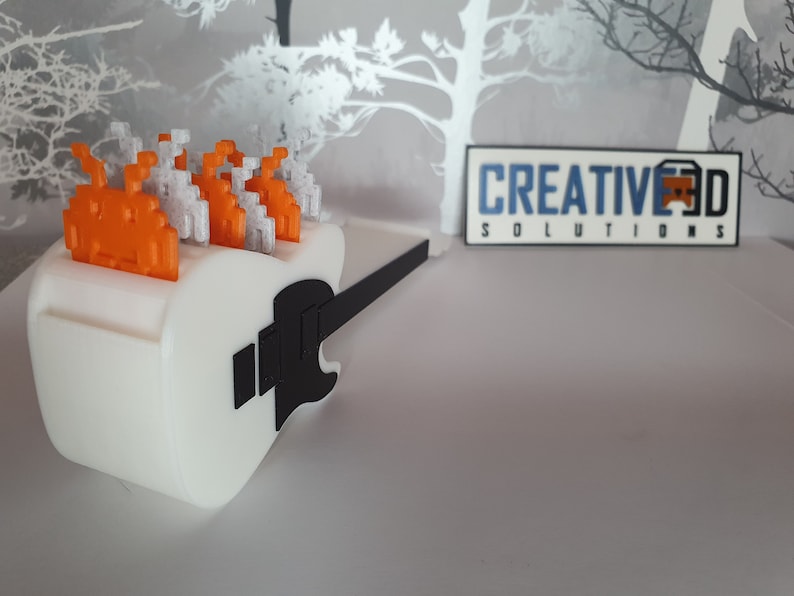 May include: A white 3D printed guitar with a black neck and a row of orange and white pixelated space invaders on top. The guitar is sitting on a gray surface. The text "CREATIVE 3D SOLUTIONS" is visible in the background.