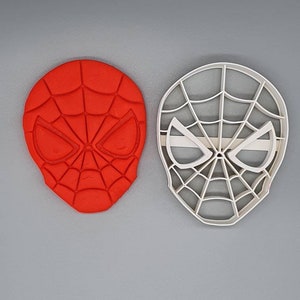 May include: A red and white plastic cookie cutter in the shape of Spider-Man's mask. The cutter is shown next to a red silicone mold of the same design.