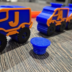 May include: A set of blue toy trucks with orange trim and letter decals. The trucks are lined up in a row on a wooden surface. The letter decals on the trucks spell out the word "PLAY".