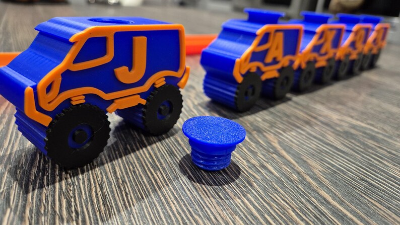 May include: A set of blue toy trucks with orange trim and letter decals. The trucks are lined up in a row on a wooden surface. The letter decals on the trucks spell out the word "PLAY".