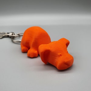 May include: Orange 3D printed dog keychain. The dog is in a laying down position with its head resting on the ground. The keychain is attached to a silver metal ring.