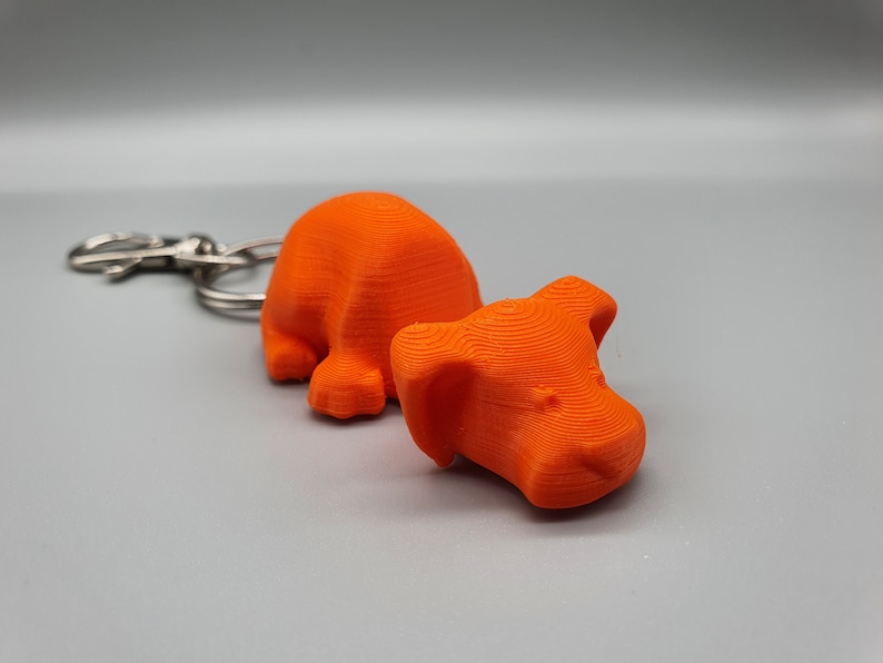 May include: Orange 3D printed dog keychain. The dog is in a laying down position with its head resting on the ground. The keychain is attached to a silver metal ring.