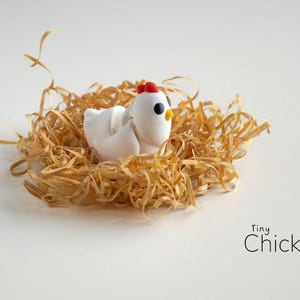 May include: A tiny white chicken figurine with a red comb and yellow beak rests in a nest of dried straw. The text "Tiny Chicken" is displayed in black script on a white background.