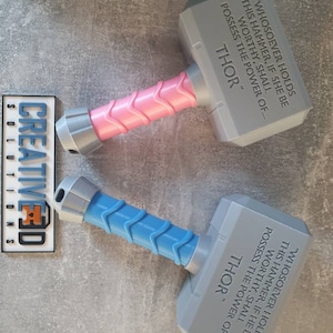 May include: Two 3D printed toy hammers with a gray head and a handle. One handle is pink and the other is blue. The hammers have the inscription "WHOSOEVER HOLDS THIS HAMMER, IF SHE BE WORTHY, SHALL POSSESS THE POWER OF... THOR" on the gray head.