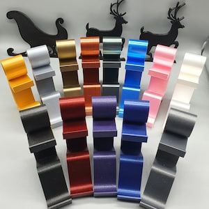 May include: A set of 14 stocking holders in various colors, including gold, silver, red, blue, black, white, and pink. The holders are shaped like a stocking and are made of a smooth, shiny material. They are arranged in a semi-circle on a gray surface.
