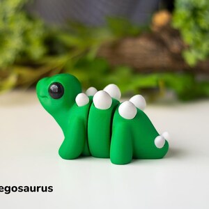 May include: A small, green, hand-painted baby stegosaurus figurine with white plates on its back. The figurine has large, black eyes and a cute, cartoonish expression.