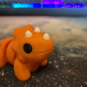 May include: A small orange dinosaur toy with white horns and black eyes. The toy is made of plastic and has a smooth, rounded surface.