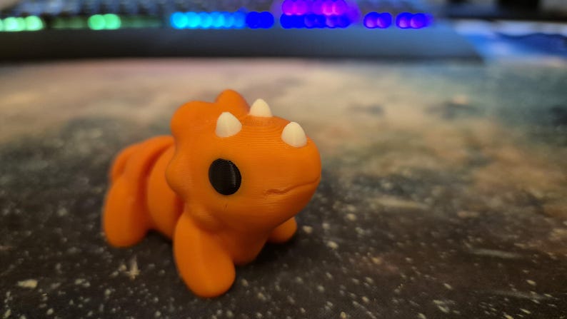 May include: A small orange dinosaur toy with white horns and black eyes. The toy is made of plastic and has a smooth, rounded surface.