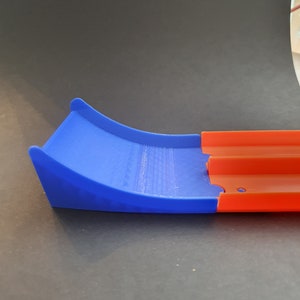 May include: A blue and orange plastic track piece for a toy car racing set. The blue piece is a curved ramp and the orange piece is a straight track section.