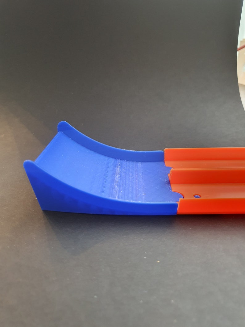 May include: A blue and orange plastic track piece for a toy car racing set. The blue piece is a curved ramp and the orange piece is a straight track section.