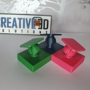 May include: Three 3D printed objects, each with a square base and a round top. The bases are green, blue, and pink. The tops are shaped like graduation caps. The objects are arranged in a triangular shape.