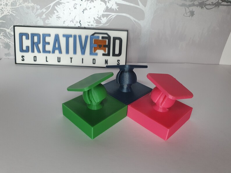 May include: Three 3D printed objects, each with a square base and a round top. The bases are green, blue, and pink. The tops are shaped like graduation caps. The objects are arranged in a triangular shape.