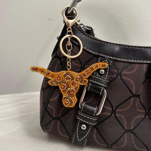 May include: A gold-colored beaded longhorn charm attached to a gold keyring. The charm is hanging from a brown and black patterned purse with a black strap and silver hardware. The charm is made of orange and gold beads.