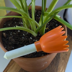 May include: A white plastic watering can with an orange plastic nozzle. The nozzle has a flower-shaped design. The watering can is sitting in a pot of green plants.