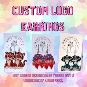 May include: Three pairs of custom logo earrings displayed on white cards. The first pair features cartoon tigers, the second pair shows red and blue Cabarrus County Fair ribbons, and the third pair depicts skull figures with the text Mariachis Cooperstown.
